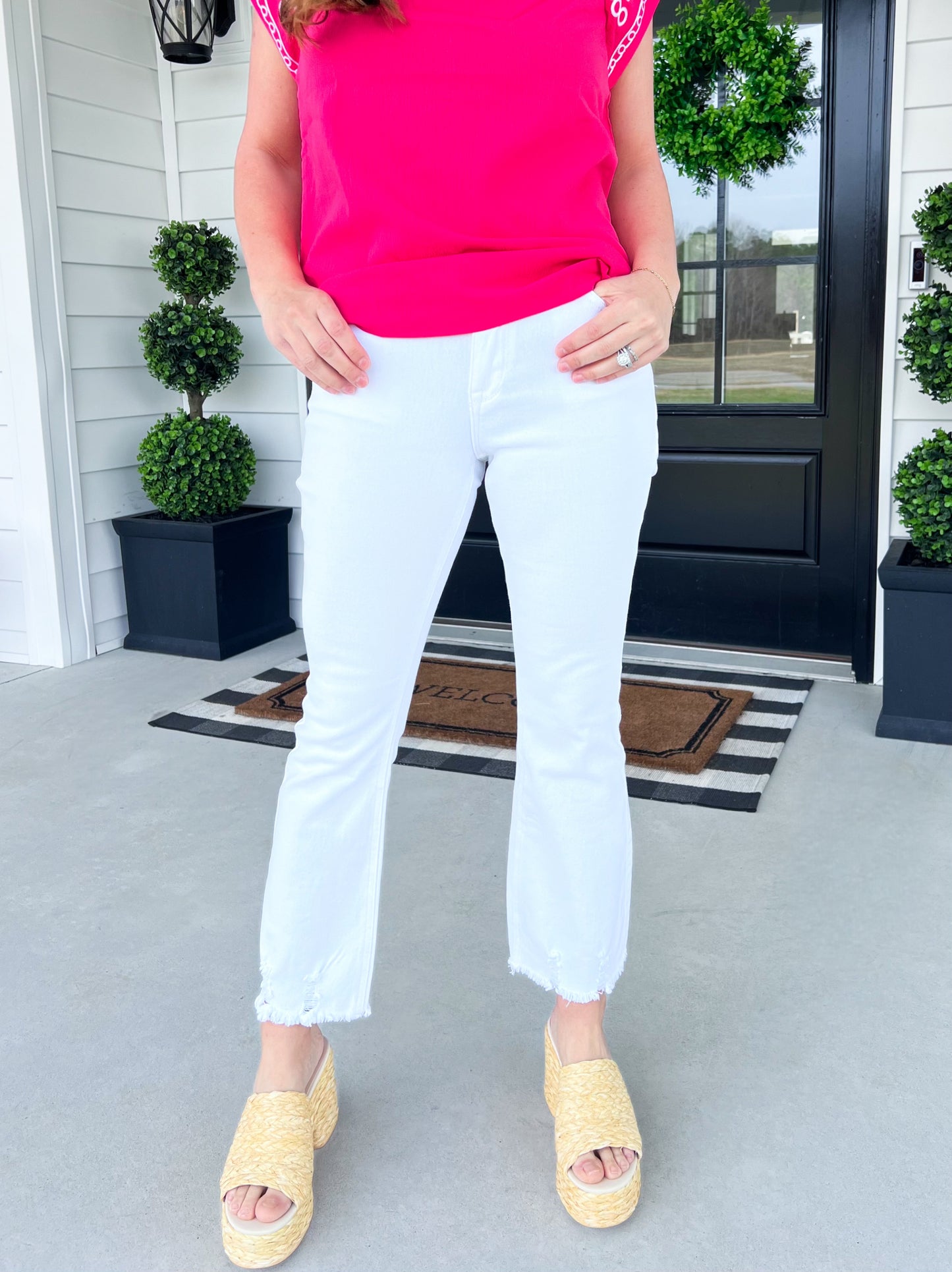 A close-up image of a model wearing white cropped flare jeans with a raw hem, paired with a bright pink sleeveless top and woven wedge sandals. The model is standing in front of a black door with greenery and a striped doormat, creating a fresh and stylish springtime aesthetic.