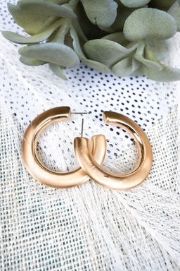 A pair of thick, gold-tone chunky hoop earrings with a modern, sculpted design, displayed against a soft fabric background.