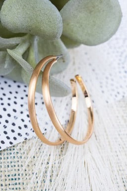 A pair of thin, gold-tone open hoop earrings with a slightly matte finish, displayed against a soft neutral backdrop.