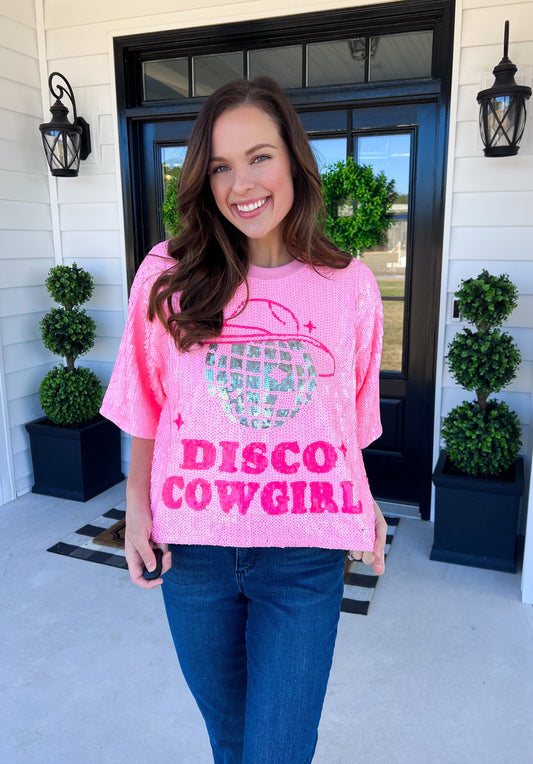 A bold pink sequin tee featuring a sparkling disco ball in the shape of a cowboy hat with "Disco Cowgirl" in vibrant lettering. Designed with a relaxed fit, short sleeves, and a fun, eye-catching shimmer. Styled with classic denim for the perfect mix of Western and party-ready vibes. Ideal for concerts, bachelorette parties, or a night out.