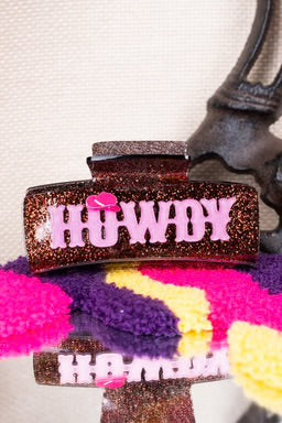  A brown glitter claw clip with "Howdy" in pink block letters and a tiny cowboy hat detail, displayed on a colorful fabric backdrop.