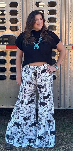 Wide-leg, high-waisted pants featuring a bold black-and-white Western print with cowboy, lasso, and rodeo-inspired illustrations. Designed with a relaxed fit and a flowy silhouette for comfort and style. Paired with a black cropped sweater and turquoise statement jewelry for a modern Western-chic look.