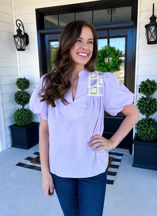 A stylish lilac blouse featuring puff sleeves, delicate lace shoulder detail, and a flattering V-neckline. Perfect for a feminine and elegant look, ideal for spring days or casual outings. Model is smiling while wearing the blouse outdoors