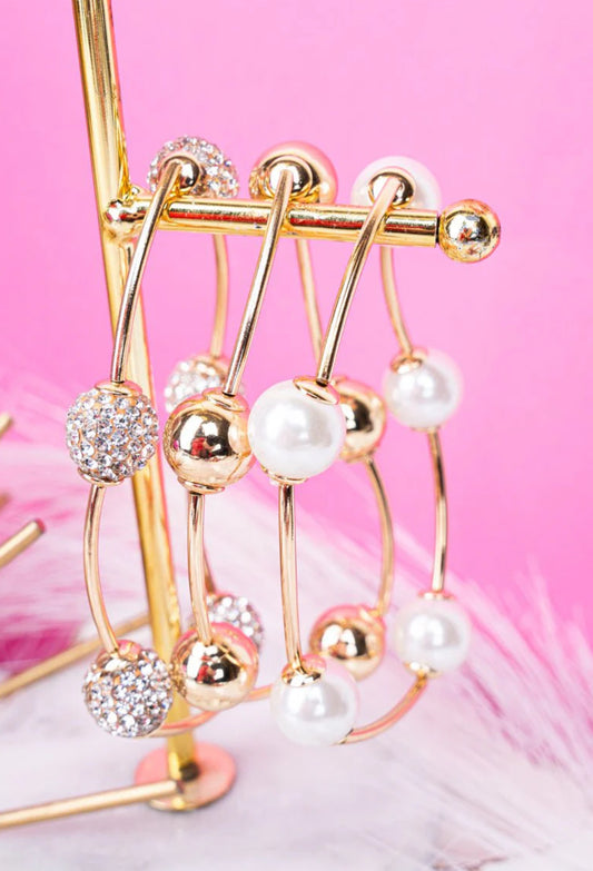  A set of gold-tone open bangles featuring pearl and crystal ball accents, displayed on a jewelry stand with a pink backdrop.