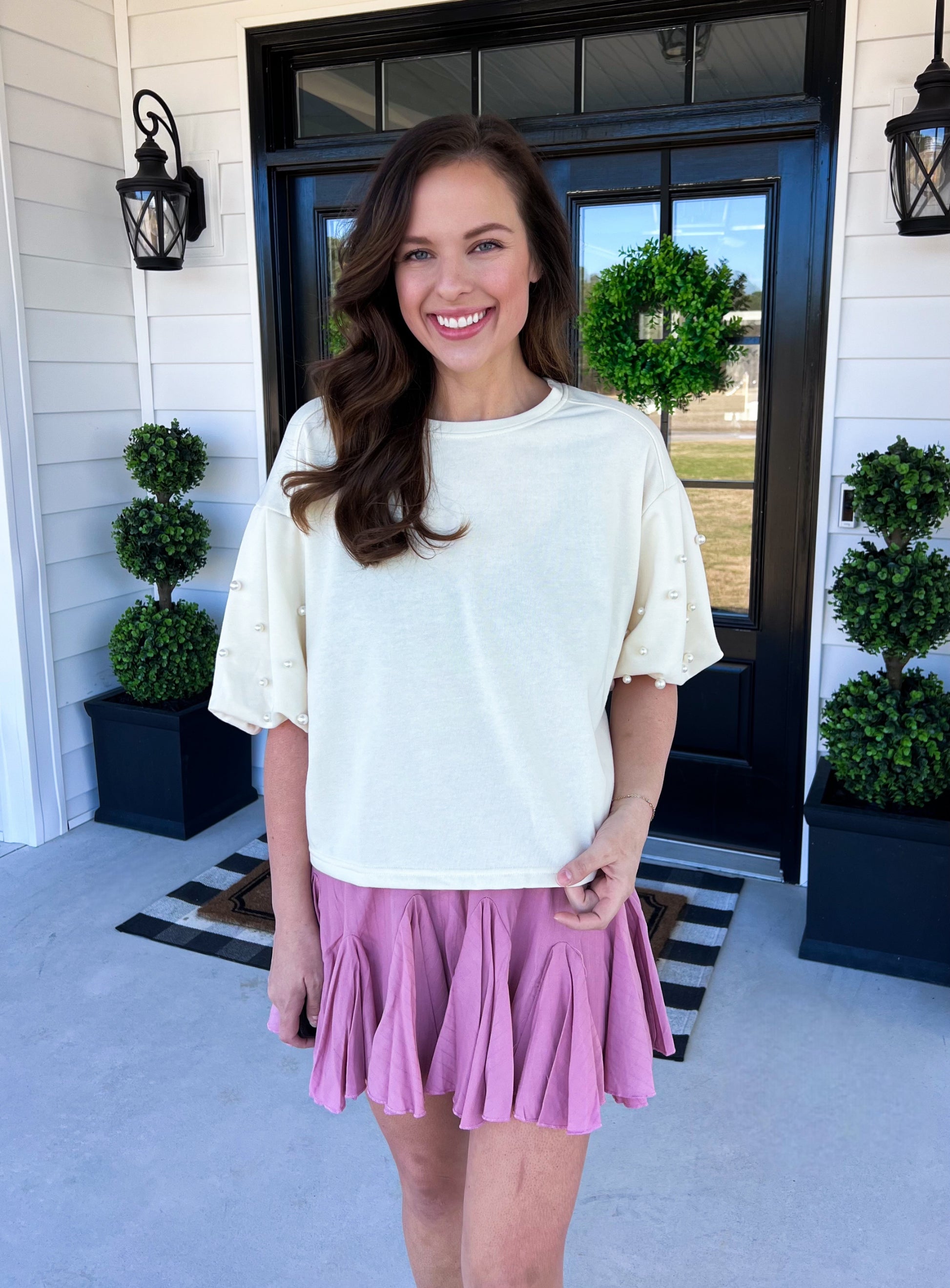 A relaxed-fit cream top featuring oversized sleeves adorned with pearl embellishments. Made from a soft, lightweight fabric with a classic crew neckline for an effortlessly chic look. Styled with a flirty mauve skort for a trendy yet comfortable outfit. Perfect for casual outings, brunch, or adding a touch of elegance to everyday wear.