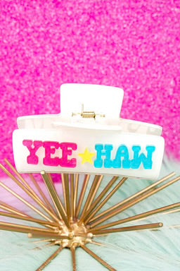  A white claw clip with "YEE HAW" in bold pink and blue lettering, displayed against a glittery pink background.