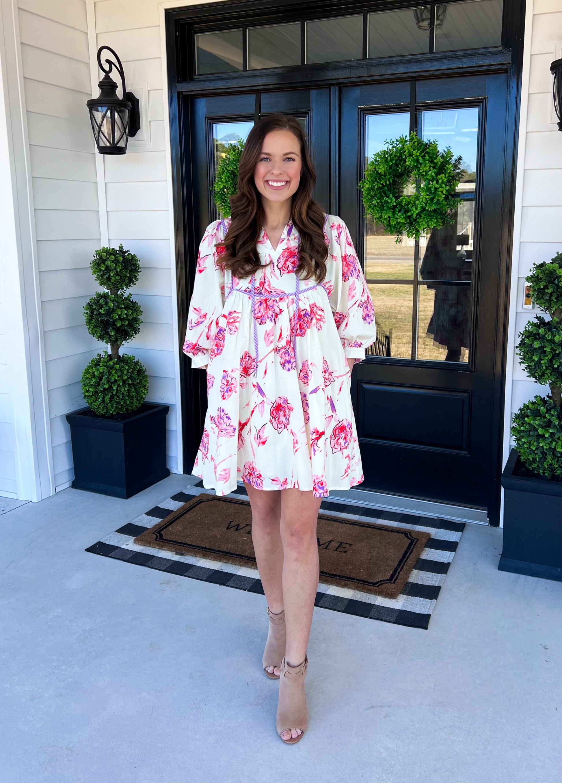 A romantic floral babydoll dress with a soft ivory base and vibrant pink rose prints. Features a flowy, tiered silhouette, billowy sleeves, and delicate lilac trim for a feminine touch. Model is smiling while wearing the dress with neutral heels on a stylish porch setting. Perfect for spring events, brunch, or a sunny day out! 