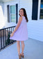 Woman looking over her shoulder and smiling while wearing a lilac V-neck sleeveless mini dress, showing the back view and flowy design of the dress.
